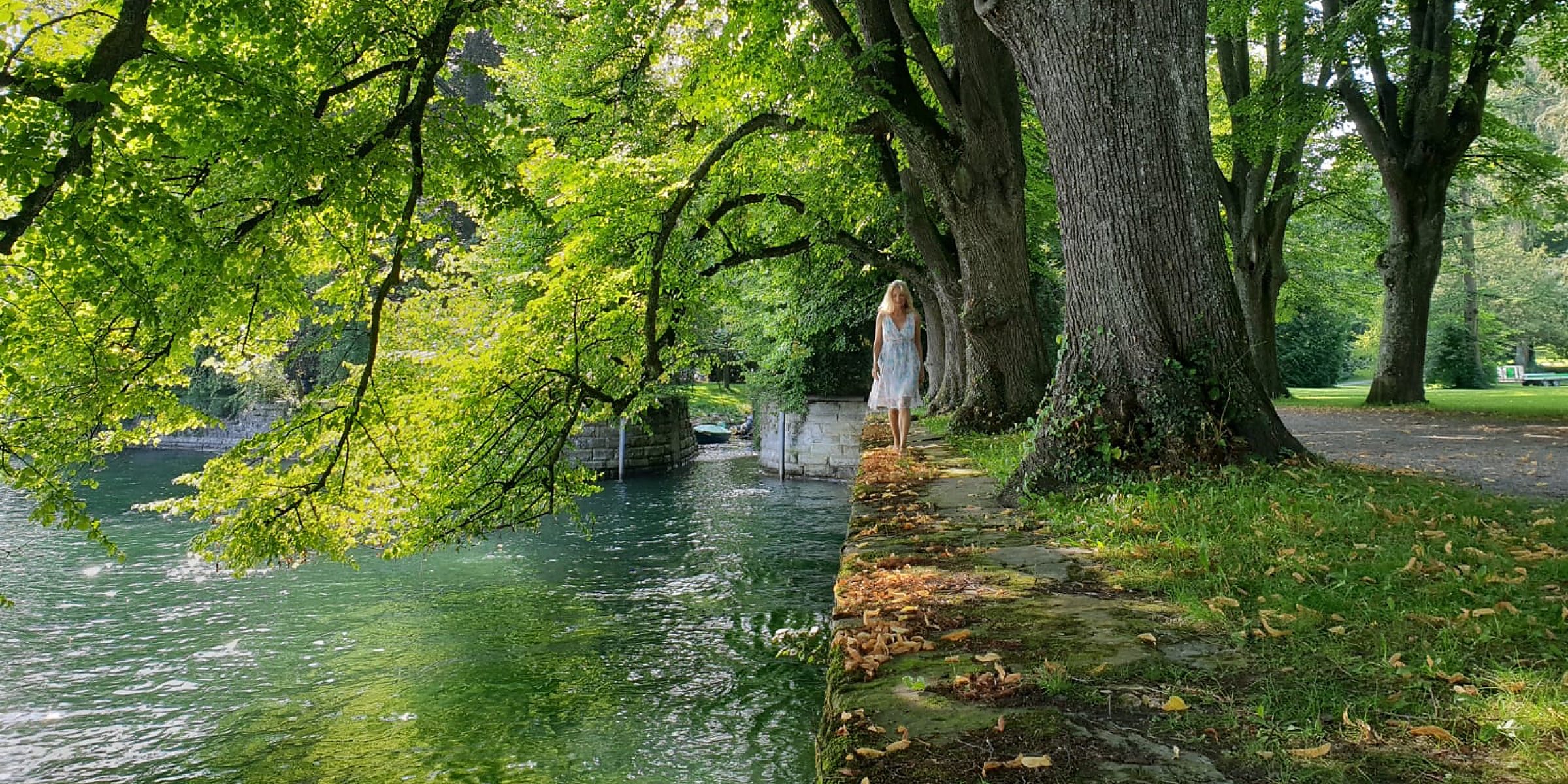Weg an einem Wasserlauf unter alten Bäumen, im Hintergrund eine Person