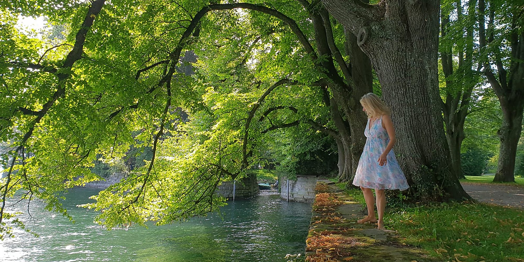 Angelika Allroggen geht barfuß unter alten Bäumen an einem Wasserlauf entlang